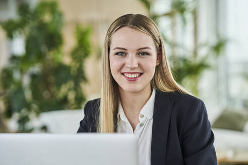 Happy blond female intern sitting in office - AKLF00518