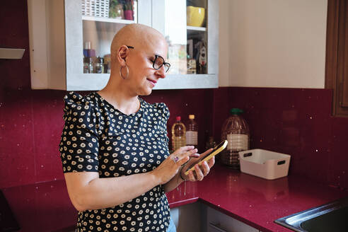Woman with cancer using mobile phone in kitchen at home - AGOF00196
