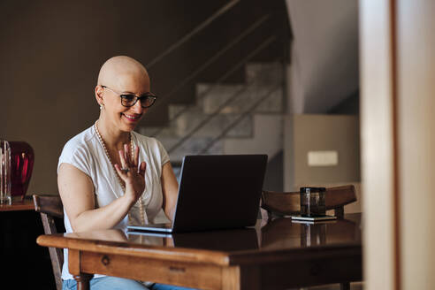 Smiling mature woman waving during video call through laptop at home - AGOF00198