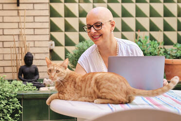 Ältere Frau mit Krebs sieht Katze an, während sie im Garten sitzt - AGOF00199