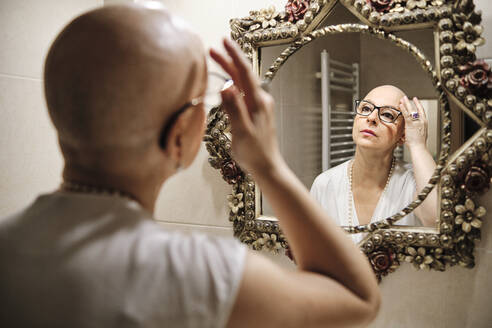 Woman with cancer touching bald head while looking at reflection in mirror - AGOF00201