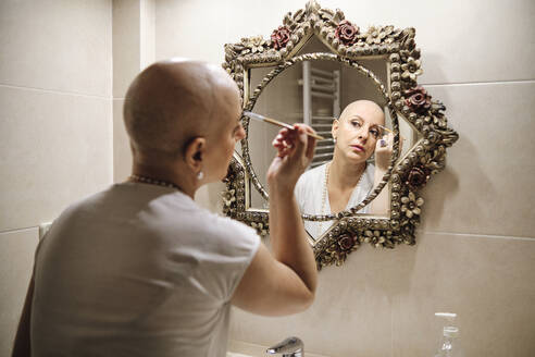 Mature woman with cancer brushing eyebrow while looking at reflection in mirror - AGOF00202
