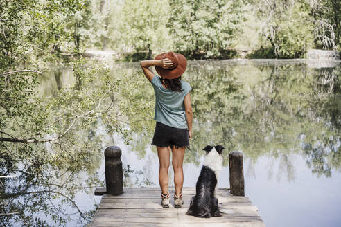 Mittlere erwachsene Frau mit Hut steht mit ihrem Hund auf einem Steg an einem See im Wald - EBBF04442