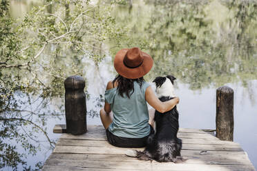 Woman wearing hat sitting with arm around pet dog on pier by lake - EBBF04443