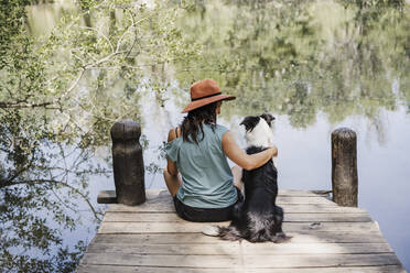 Mid adult woman sitting with Border Collie on pier by lake in forest - EBBF04444