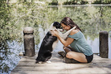 Smiling woman embracing dog while sitting on pier by lake in forest - EBBF04445