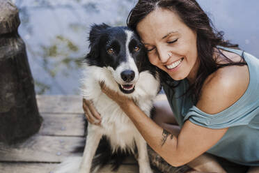 Lächelnde Frau mit geschlossenen Augen, die ihren Hund umarmt, während sie auf einem Steg sitzt - EBBF04448
