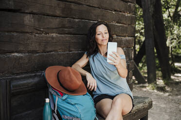 Mittlere erwachsene Frau, die ein Selfie mit ihrem Smartphone macht, während sie vor einer Hütte im Wald sitzt - EBBF04449