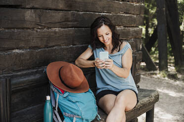 Lächelnde Frau, die auf einer Bank im Wald sitzt und über ihr Mobiltelefon Textnachrichten verschickt - EBBF04450