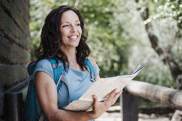 Lächelnde Touristin schaut weg, während sie eine Karte im Wald hält - EBBF04456