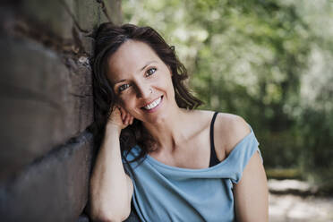 Happy mid adult woman leaning on cottage in forest - EBBF04459