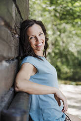 Happy woman leaning on cottage while standing in forest - EBBF04463