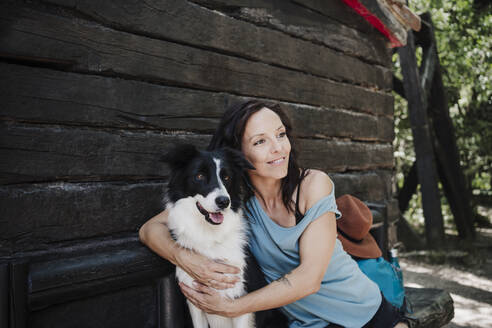 Mid erwachsene Frau sitzt mit Arm um Hund außerhalb Hütte im Wald - EBBF04489