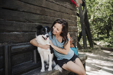 Happy woman looking at pet dog while sitting with arm around in front of cottage - EBBF04490