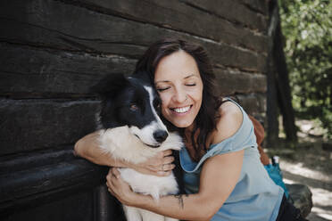 Lächelnde erwachsene Frau mit geschlossenen Augen, die einen Border Collie vor einem Haus umarmt - EBBF04491