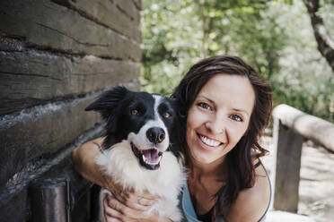 Happy woman sitting with arm around pet dog by cottage in forest - EBBF04497