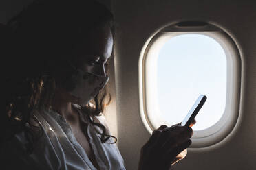 Female professional using smart phone in airplane during pandemic - JAQF00697