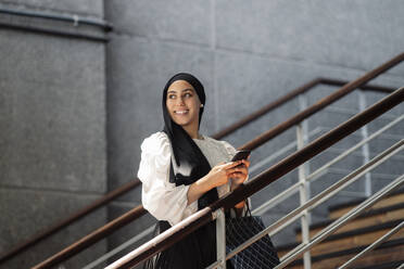 Lächelnde junge Frau mit Mobiltelefon, die auf einer Treppe steht und wegschaut - JCCMF03427