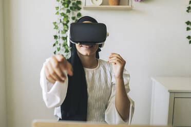 Smiling female freelance worker pointing while using virtual reality headset at home - JCCMF03455