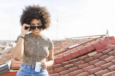 Fröhliche junge Afro-Frau mit Einwegbecher und Sonnenbrille auf dem Dach - IFRF00975