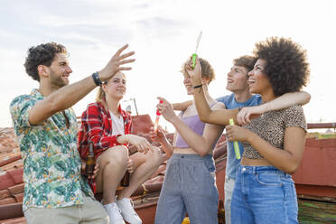 Cheerful multi-ethnic male and female friends playing with bubbles on rooftop - IFRF01020