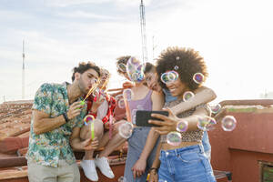 Young male and female friends taking selfie while blowing bubbles on terrace - IFRF01023