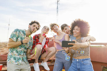 Cheerful multi-ethnic friends taking selfie while playing with bubbles on terrace - IFRF01024