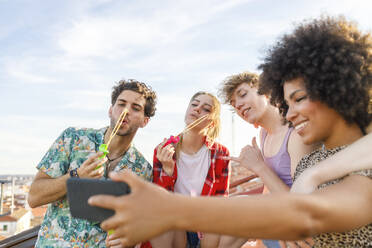 Young male and female friends blowing bubbles while taking selfie on rooftop - IFRF01025