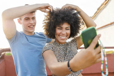 Fröhliches junges Paar, das ein Selfie mit seinem Smartphone macht, während es auf dem Dach mit seinen Haaren spielt - IFRF01026