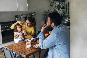 Frau füttert ihre Tochter mit Essen, während sie mit einem Mann am Tisch sitzt - ASGF01012