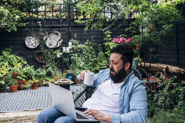 Mann mit Kaffeetasse und Laptop im Hinterhof sitzend - ASGF01014