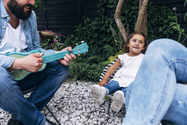 Lächelndes Mädchen, das seine Mutter anschaut, während der Vater im Hinterhof Gitarre spielt - ASGF01020