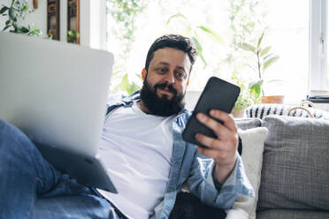 Mann mit Laptop, der ein Mobiltelefon benutzt, während er im Wohnzimmer sitzt - ASGF01031