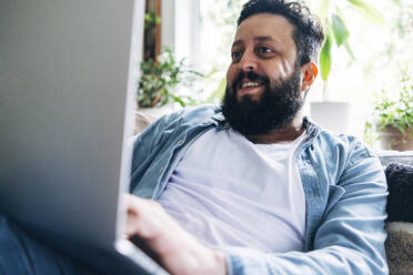 Lächelnder bärtiger Mann mit Laptop bei der Arbeit zu Hause - ASGF01032