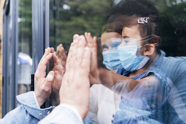 Familie mit Gesichtsschutz grüßt durch eine Glastür während COVID-19 - ASGF01046