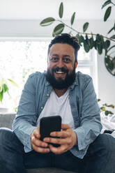 Lächelnder Mann mit Mobiltelefon in der Hand, der zu Hause sitzt - ASGF01059