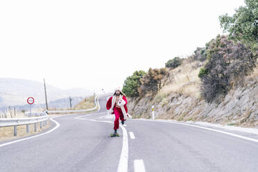 Junger Mann im Weihnachtsmannkostüm fährt auf der Straße Skateboard - OCMF02197