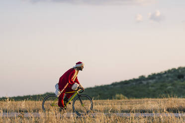 Junger Mann im Weihnachtsmannkostüm fährt bei Sonnenuntergang Fahrrad auf der Straße - OCMF02202