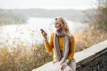 Lächelnde blonde Frau, die während eines Videogesprächs über ein Mobiltelefon auf einer Stützmauer sitzt - JOSEF05342