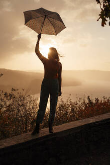 Silhouette of woman with umbrella standing on retaining wall at sunset - JOSEF05352
