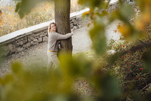 Woman with eyes closed embracing tree during autumn - JOSEF05358