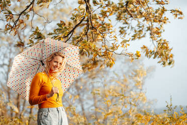 Lächelnde Frau mit gepunktetem Regenschirm an einem sonnigen Tag - JOSEF05372