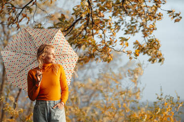 Frau schaut weg, während sie mit der Hand in der Tasche steht und einen Regenschirm an einem sonnigen Tag hält - JOSEF05373