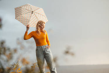 Lächelnde blonde Frau hält einen Regenschirm, während sie mit der Hand in der Tasche steht - JOSEF05374