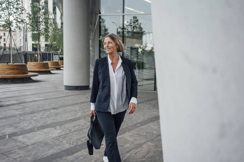 Smiling female professional with bag looking away while walking on walkway - VPIF04455