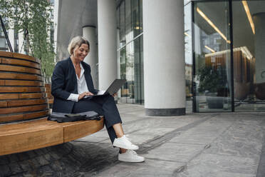 Lächelnde Geschäftsfrau, die einen Laptop benutzt und auf einer Bank im Büropark sitzt - VPIF04456