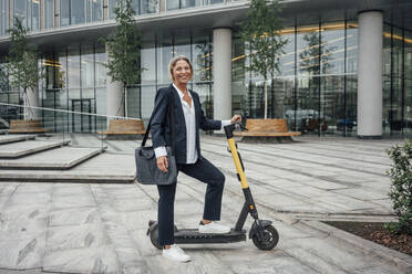 Female professional smiling while standing with electric push scooter at office park - VPIF04458