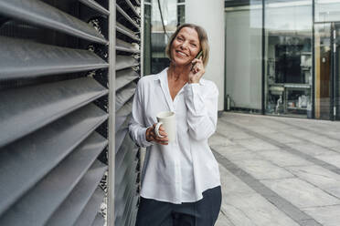 Geschäftsfrau mit Tasse, die mit ihrem Smartphone telefoniert, während sie an den Jalousien im Büropark steht - VPIF04463