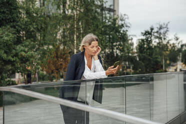 Mature businesswoman using smart phone while leaning on glass railing - VPIF04472