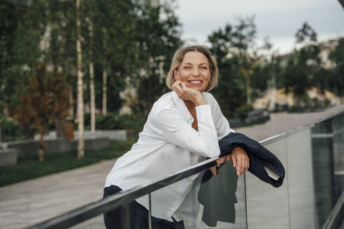 Smiling mature businesswoman with hand on chin leaning on railing - VPIF04473